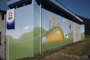 Vue de la centrale hydroélectrique dont la façade est entièrement peinte d’un paysage onirique : ciel azur, ampoules oranges sur les collines vertes et rondes sur lesquelles paissent des moutons.
