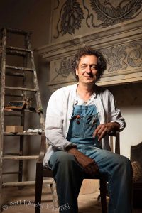 Portrait assis de Thierry Battaglia en tenue de travail (salopette en jean), pris dans un rayon de lumière devant un décor en cours de restauration.